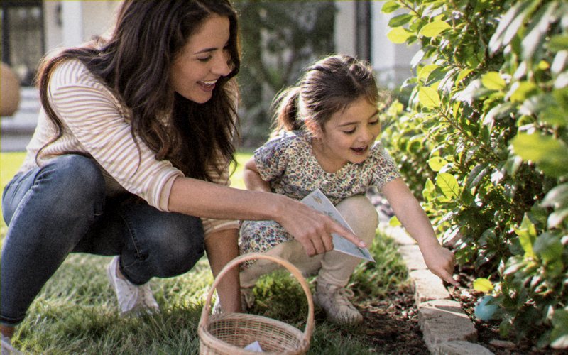 A mother and her young daughter crouching by a garden border, one holding a clue card and the other pointing at something hidden.