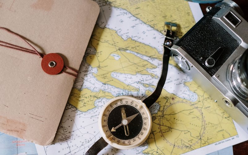 Old maps spread over a table with a compass, a camera and a notebook tied with string.