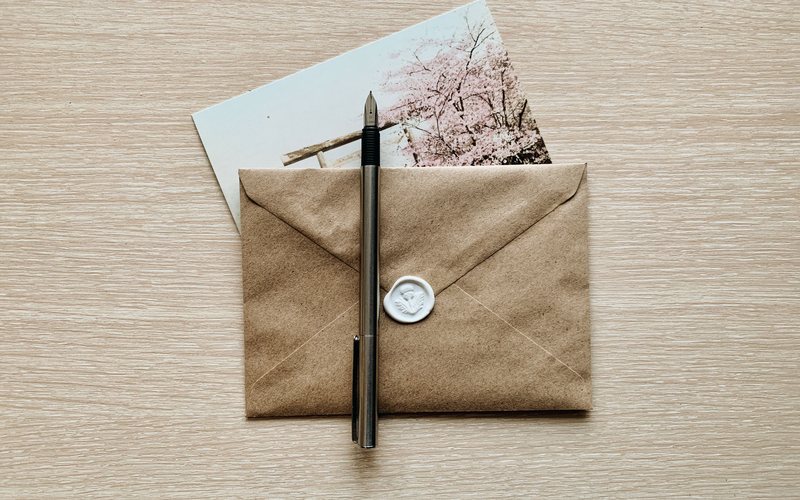 A brown envelope closed with a white wax seal, a pen lying across it on a wooden desk.