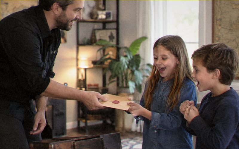 An adult handing a wax-sealed envelope to two delighted children, an open chest beside them.
