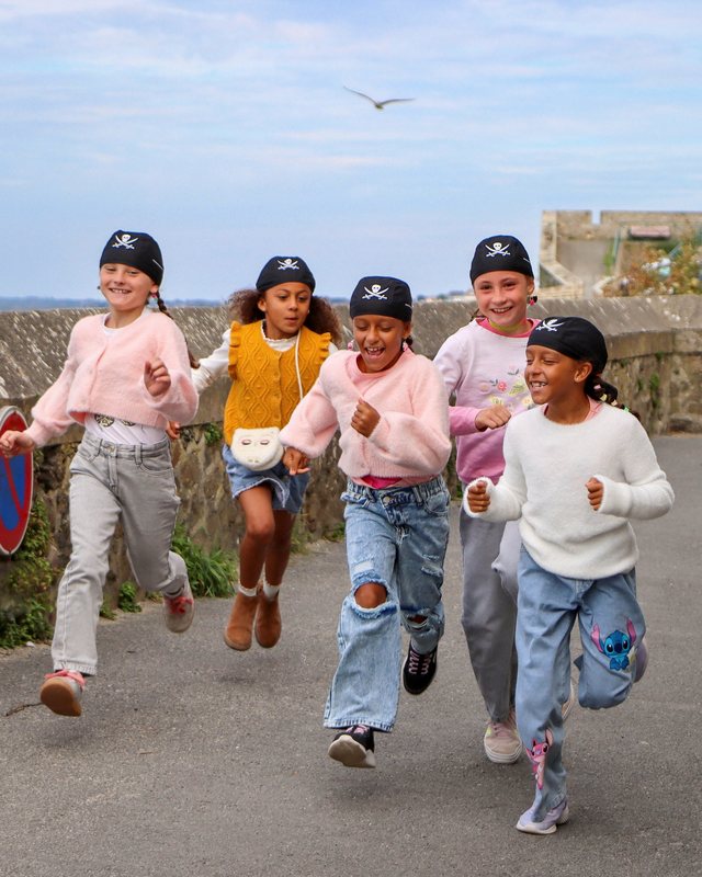 Five children in pirate bandanas running and laughing along a seaside lane.